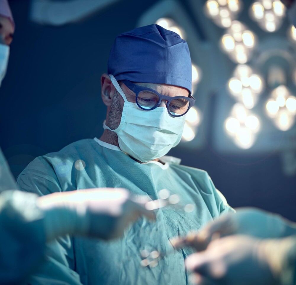 Herr Dr. Lautersack in blauer OP-Bekleidung mit Deckenbeleuchtung im Hintergrund. Im Vordergrund wird Werkzeug weitergereicht.